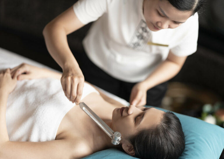 A spa therapist uses a tuning fork to relax a guest laying on a massage bed by placing the vibrations next to her ear