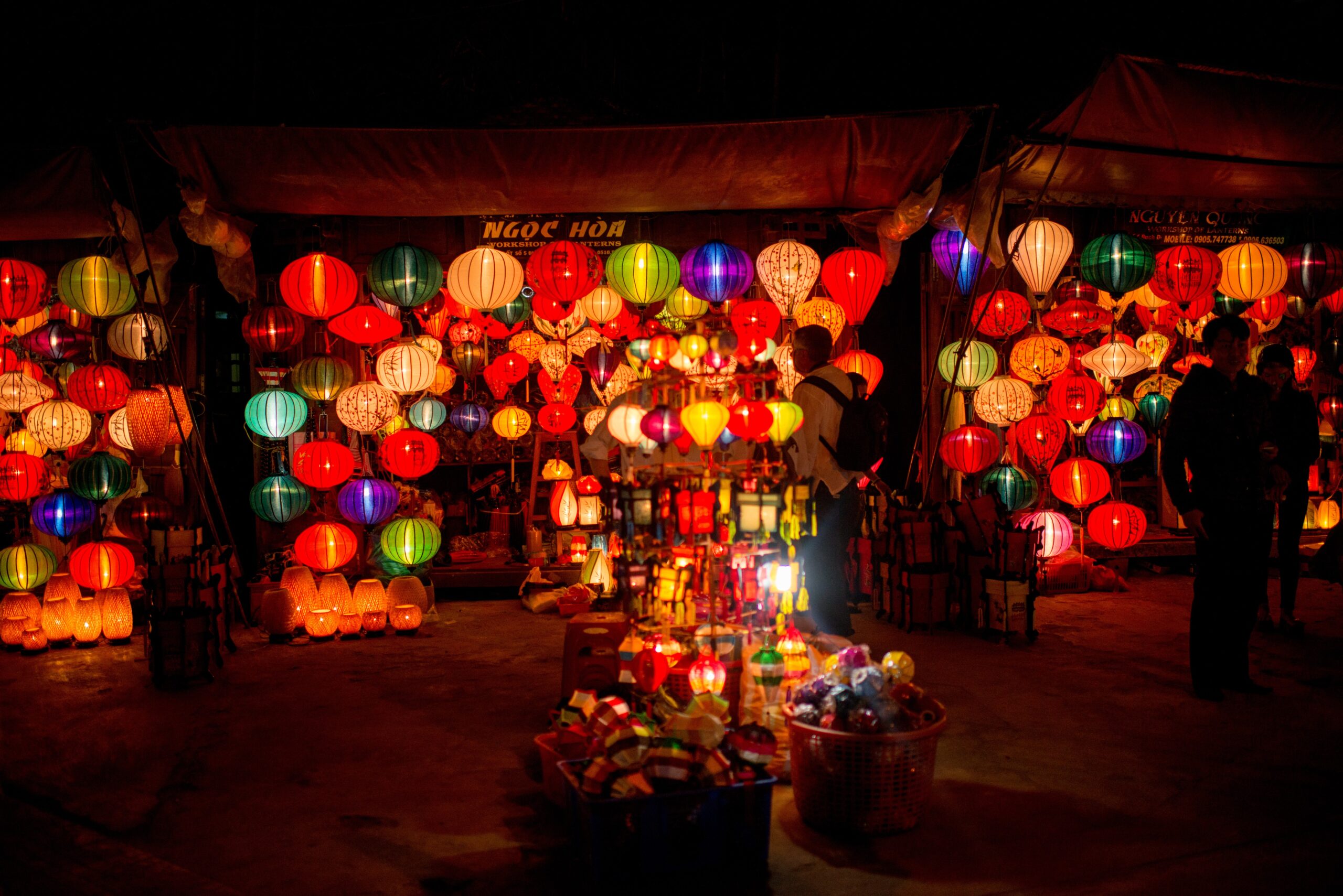 A man stoped and looks at a lantern stall at night, the whole street side is lit with hundreds of different style lanterns with many colours that set the street and surrounding aglow.