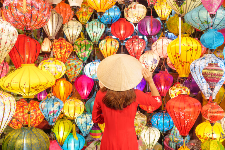 happy woman wearing Ao Dai Vietnamese dress with looks out at a wall of colorful lanterns for sale